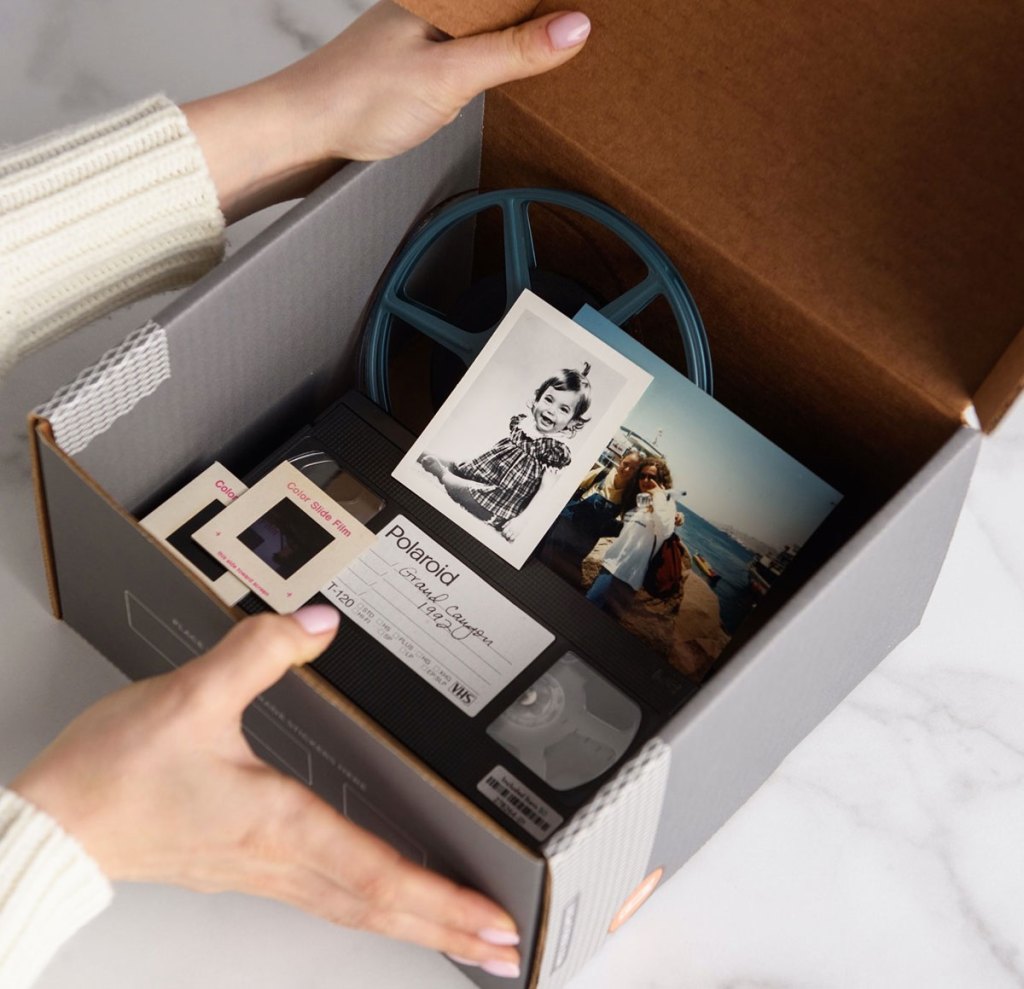 woman holding a square cardboard box with old photos, a film reel, and vhs tape inside