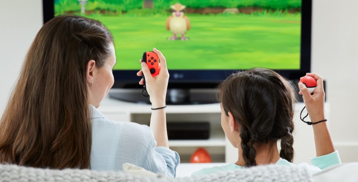 woman and girl sitting on couch playing game with Nintendo Switch gaming console and Poke Ball controllers on TV