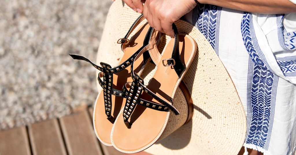 women in white and blue dress holding sun hat and pair of black leather studded sandals in her hands