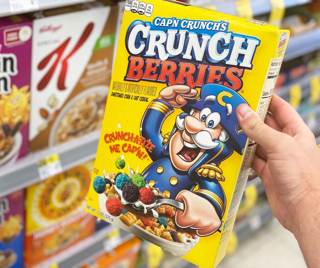 person holding a yellow box of capn crunch berries cereal in front on shelves of cereal