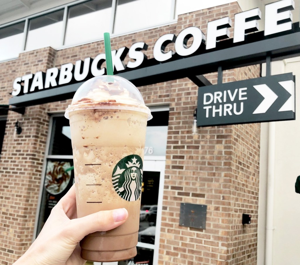 person holding up frappuccino drink in front os starbucks drive-thru sign