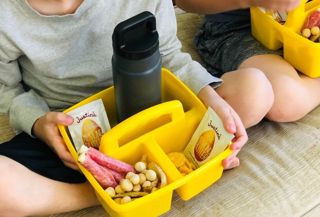 kid holding a yellow caddy with snacks and water bottle inside