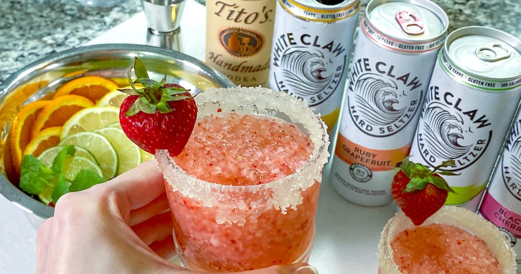 hand holding white pink drink with strawberry and white claw cans in background