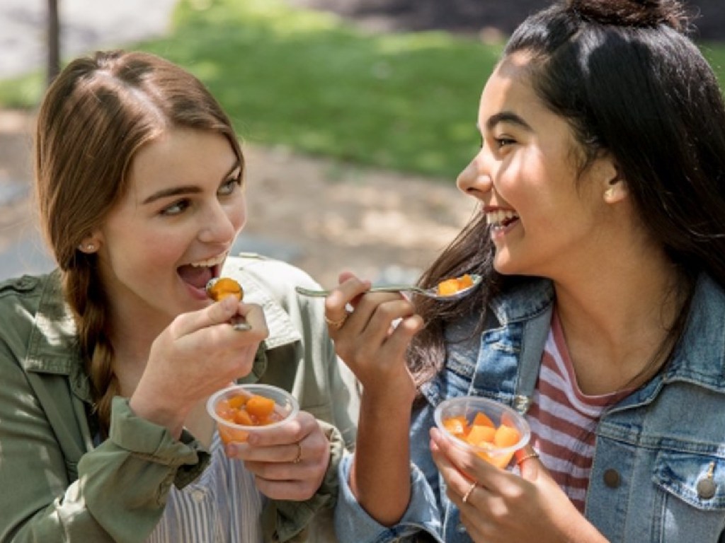 2 girls facing each other eating del monte fruit cups while smiling