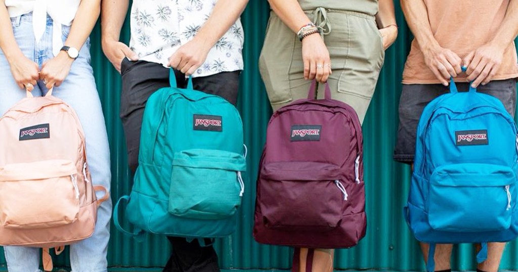 people standing against green wall holding jansport brand backpacks in front of them