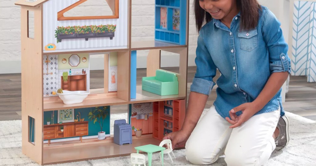 girl kneeling on floor next to wooden dollhouse playing with doll table and chair set