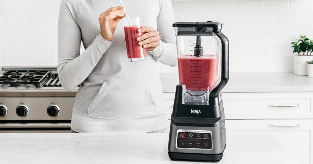 woman holding red smoothie standing next to kitchen counter with ninja blender