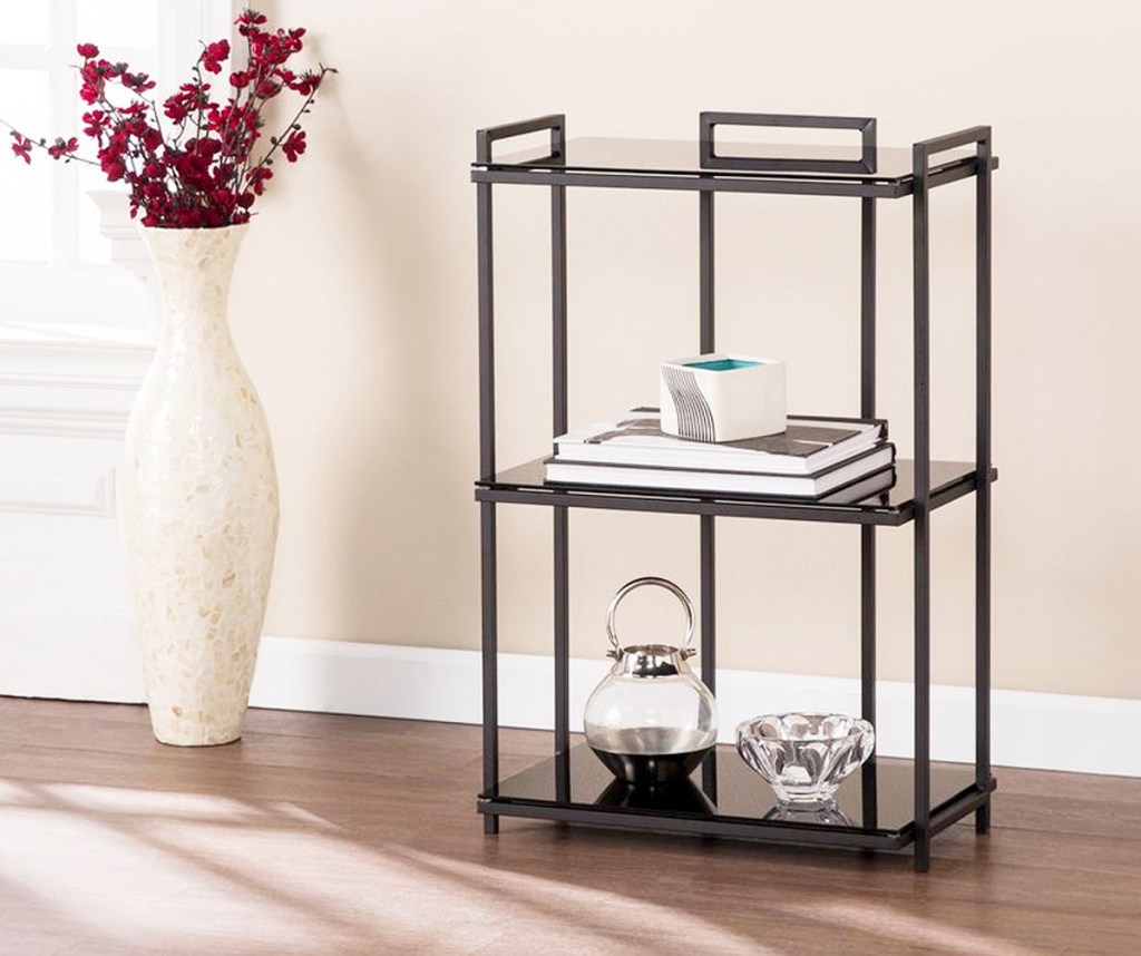 three tier glass bookcase with black metal frame on hardwood floor near large vase of red flowers