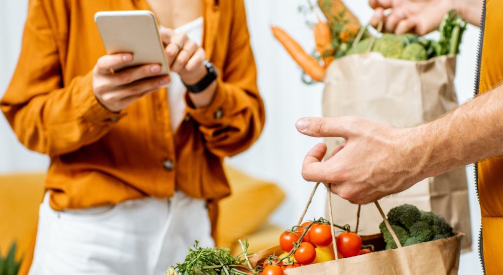 person holding phone with other persons hands holding brown bags of groceries