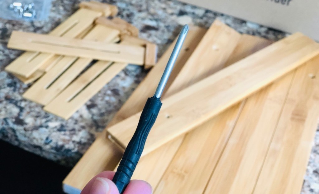 Hand holding a black and silver mini Phillips head screwdriver with wood pieces on countertop in background