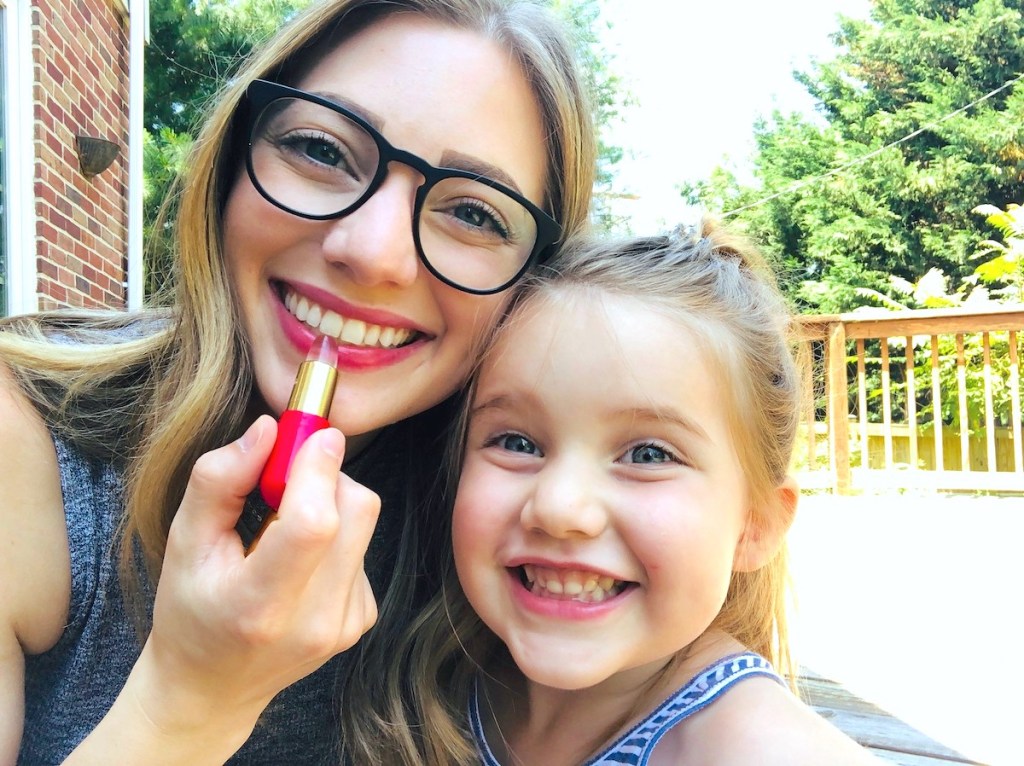 woman applying lipstick with smiling girl next to her