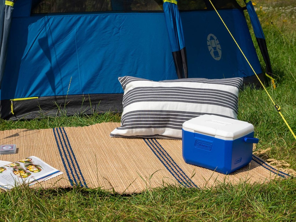 blue coleman cooler sitting on a blanket next to a pillow and magazine outside of a blue tent