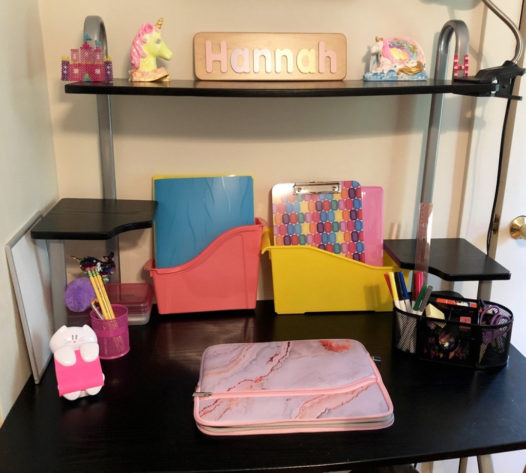 black desk with pink laptop case, post-it note dispenser, plastic bins of workbooks, and pens and pencils