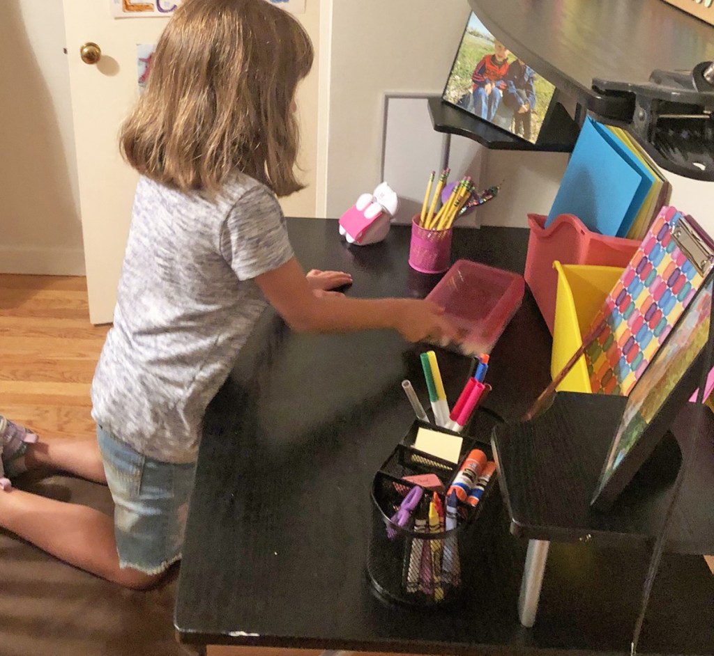 girl with brown hair kneeling on chair at desk reaching into pencil box on a black desk full of school supplies