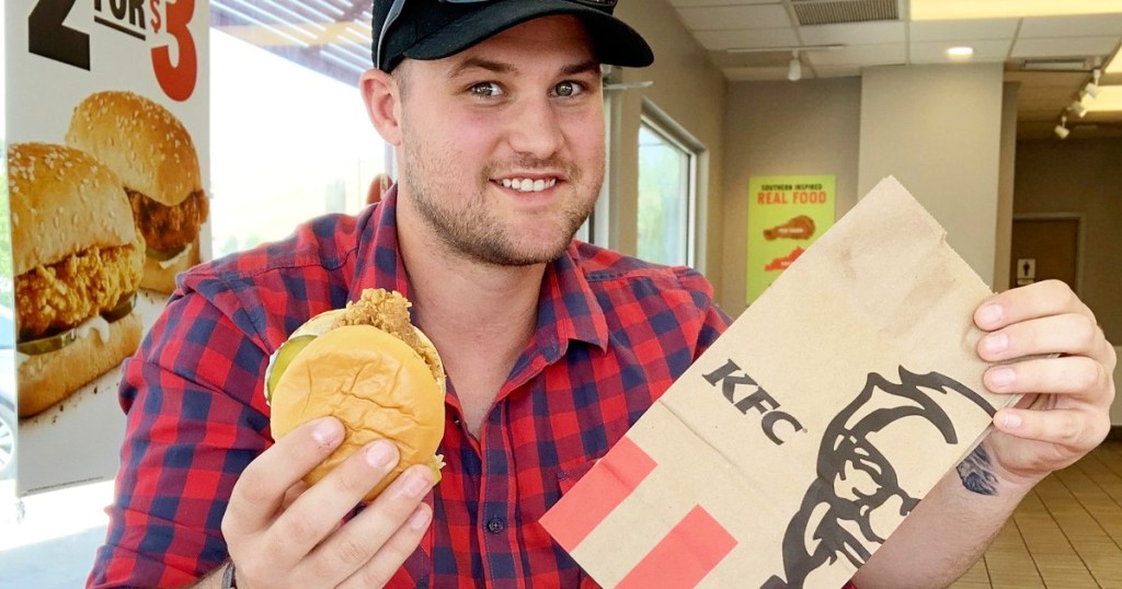 man in red and black plaid shirt holding up a brown KFC bag and fried chicken sandwich