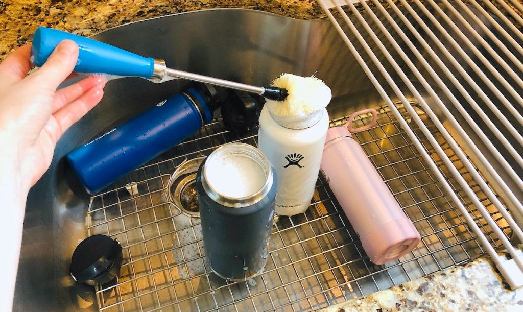 hand holding a blue bottle brush cleaning various water bottles in stainless steel sink