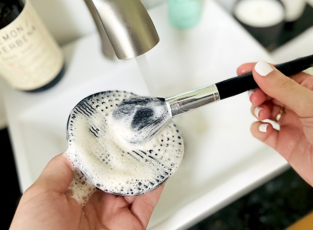 hands cleaning makeup brush on black silicone mat over bathroom sink