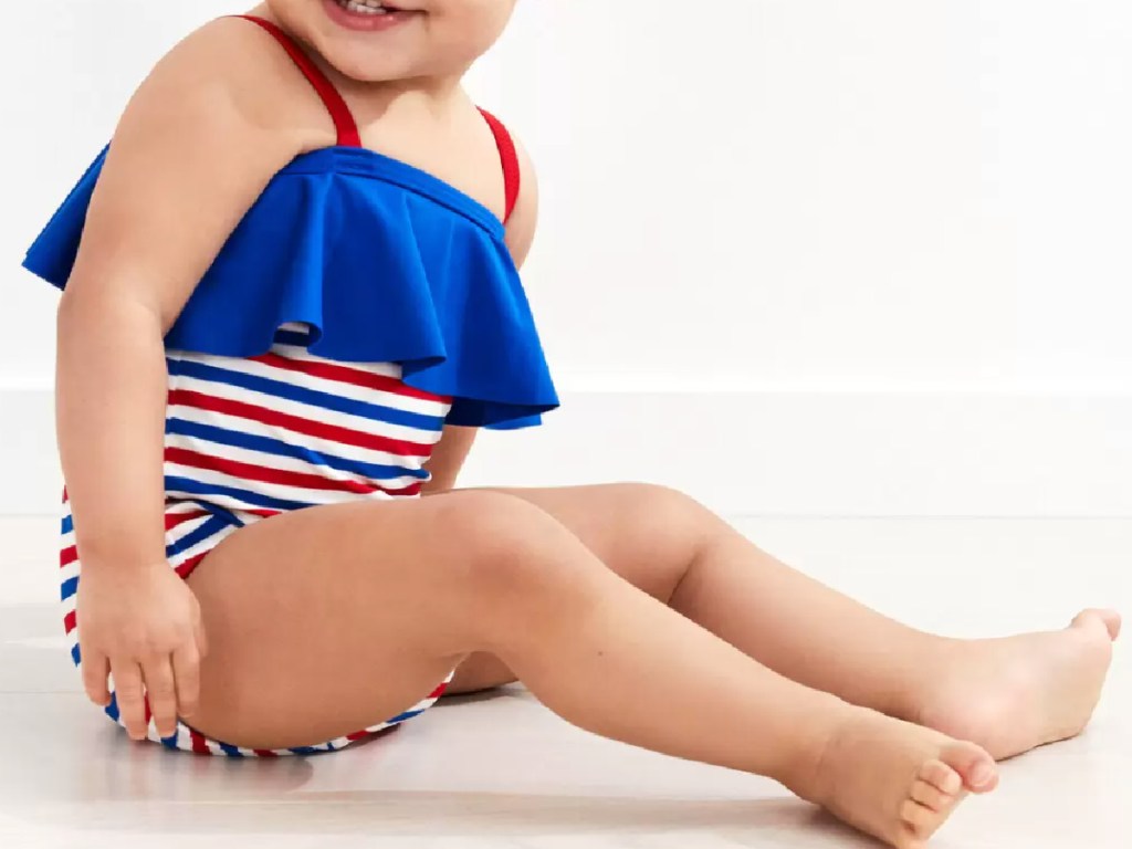 infant baby girl sitting sideways wearing a red, white and blue striped swimsuit