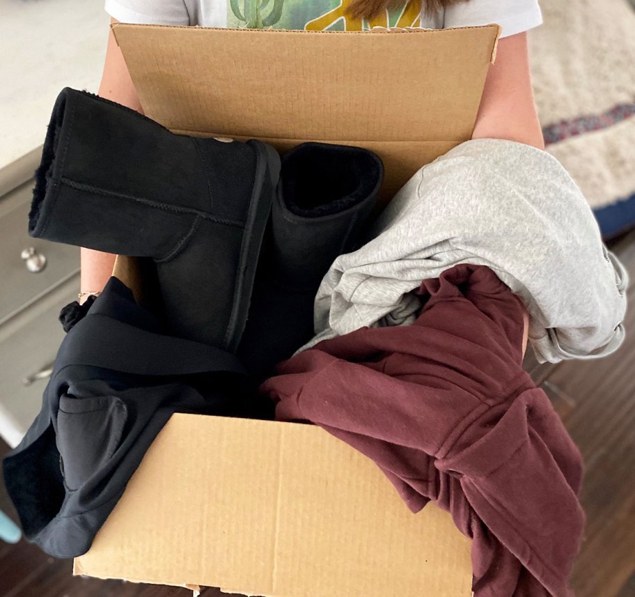 person holding up a brown shipping box full of pieces of clothing and pair of black boots