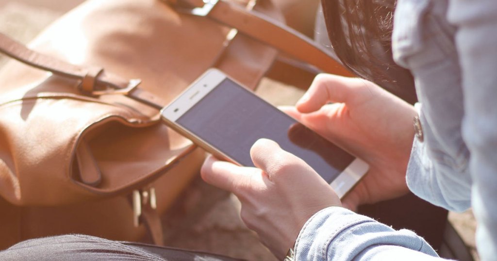 woman sitting holding a white smartphone near a leather purse