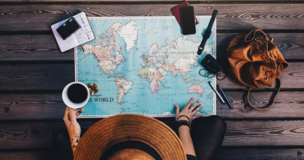 person wearing brown hat looking down at paper world map with cup of coffee