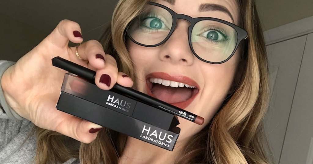 woman with blonde hair and black glasses holding up haus laboratories liquid lip gloss and lip pencil