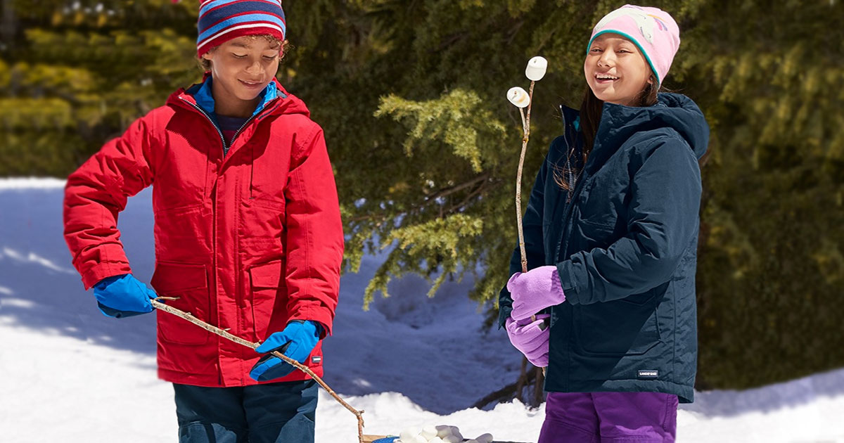two kids wearing winter jackets and accessories while in snow roasting marshmallows