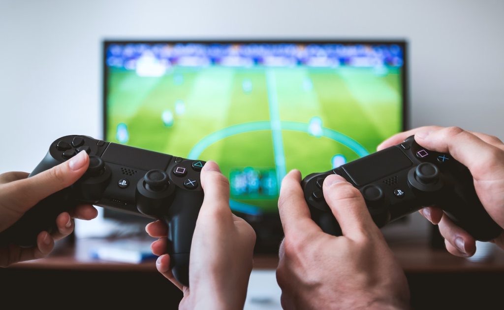 two hands holding gaming remote controls in front of football tv screen