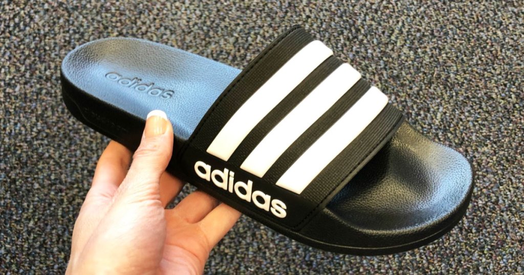 person holding up a black adidas slide sandal with three white stripes across the top