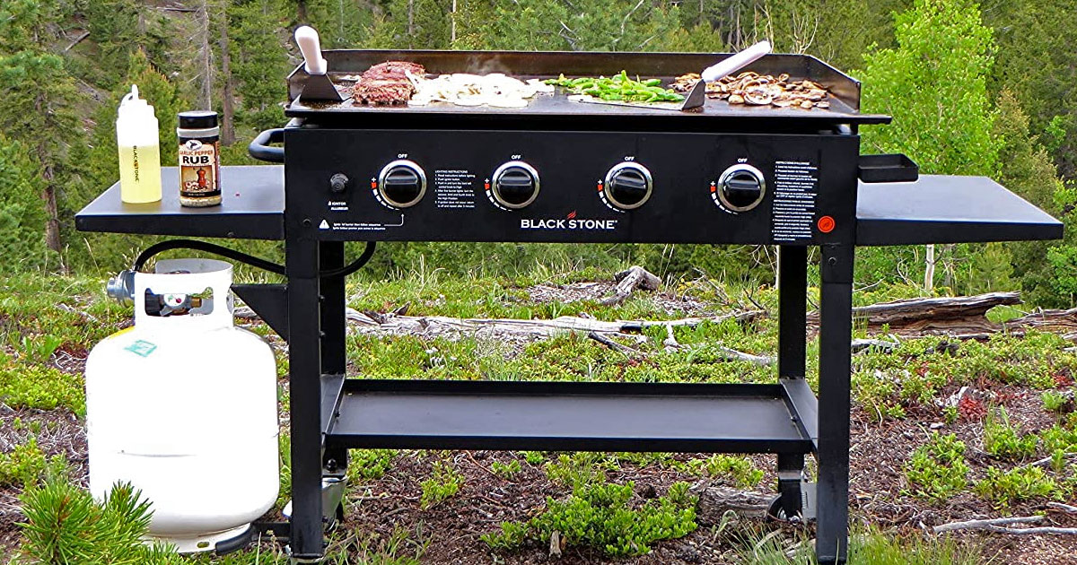 black blackstone griddle with food on the griddle and accessories on side shelf and white propane tank hooked up to it