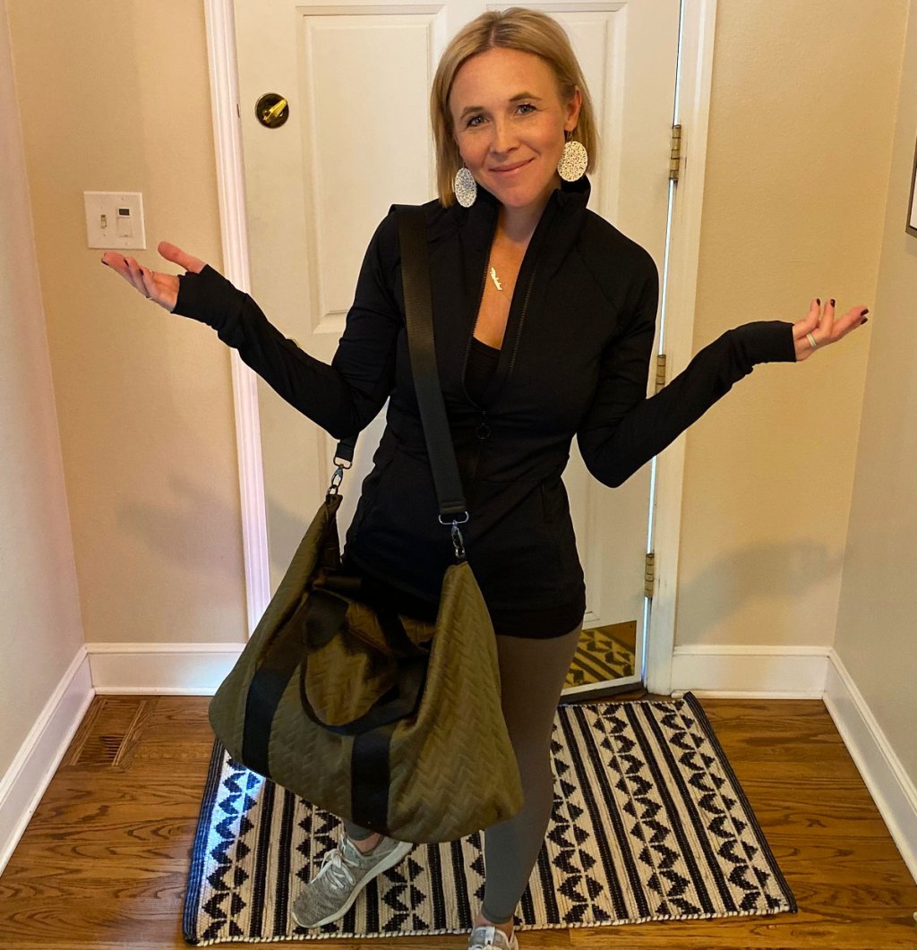 woman with blonde hair standing in entryway wearing black jacket, grey leggings, and green gym bag on shoulder