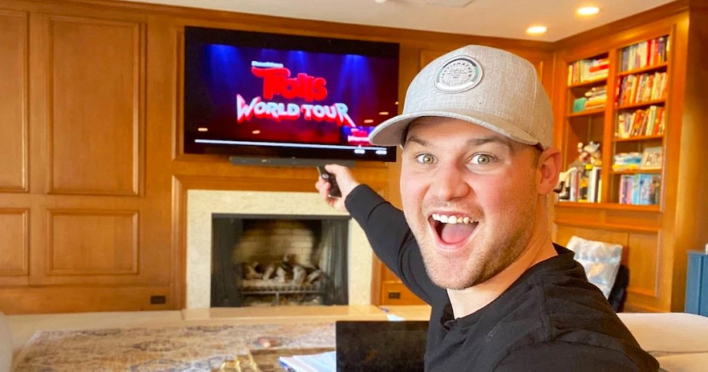 man wearing grey hat and black shirt pointing remote at TV watching Trolls World Tour movie