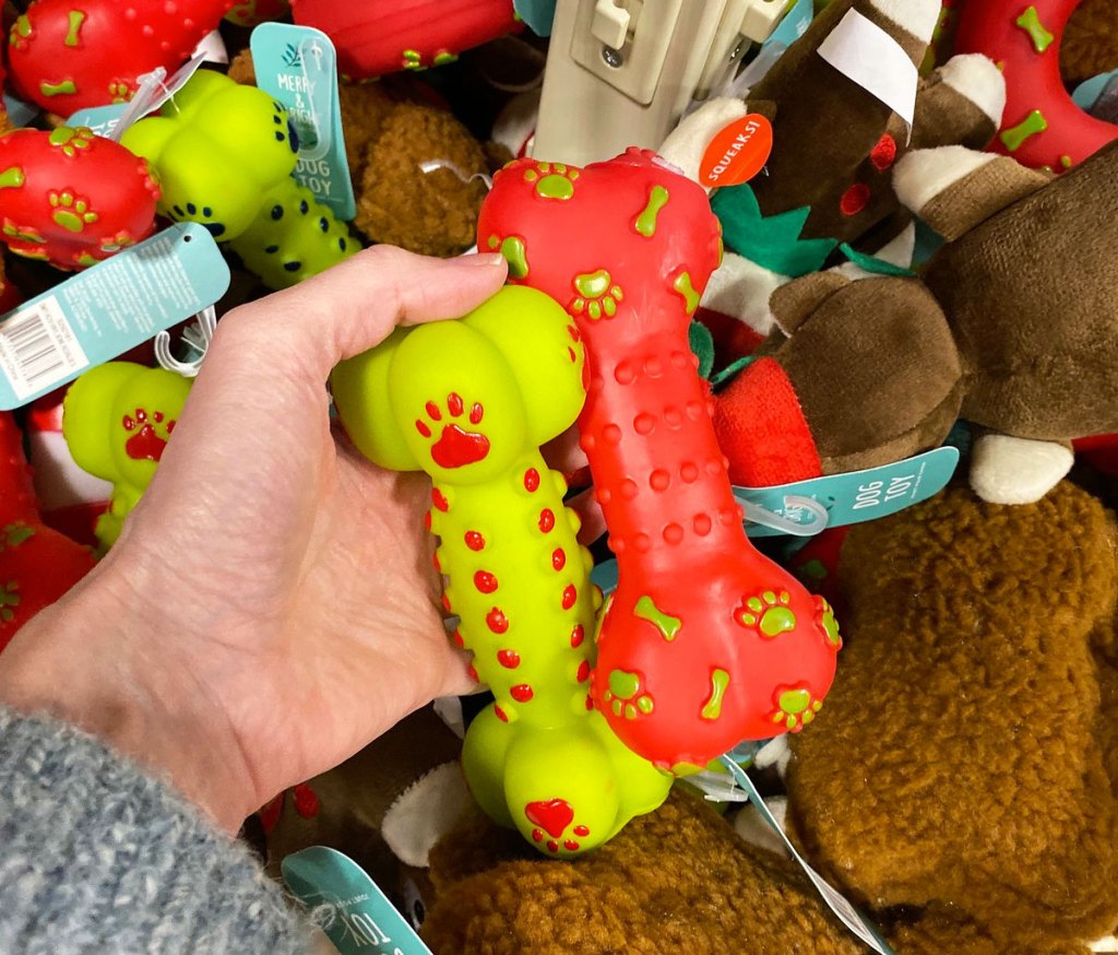 woman in grey sweater holding up green and red plastic dog bone toys in front of other christmas toys