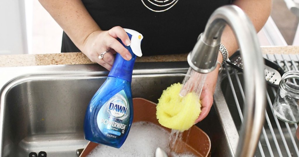 woman holding blue bottle of dawn powerwash soap spray and yellow scrub daddy sponge in kitchen sink
