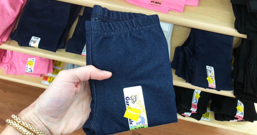 woman holding up a pair of denim blue baby girls leggings with shelf of leggings in background