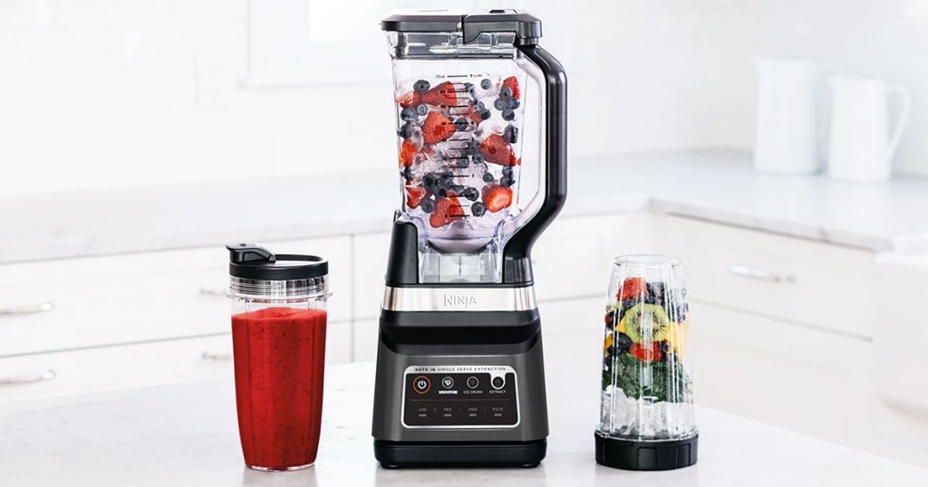 ninja blender with fruit inside on a kitchen counter with two individual blender cups next to it