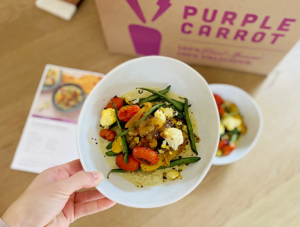 hand holding bowl of cooked vegetables with purple carrot box