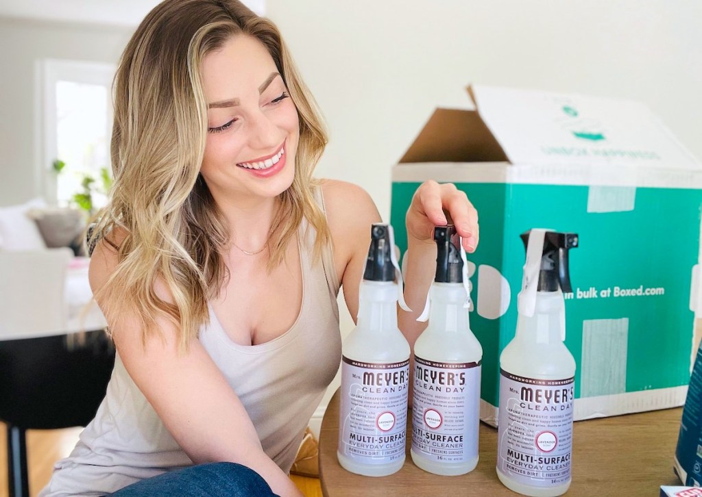 woman sitting at table next to three bottles of mrs meyers lavender cleaning spray