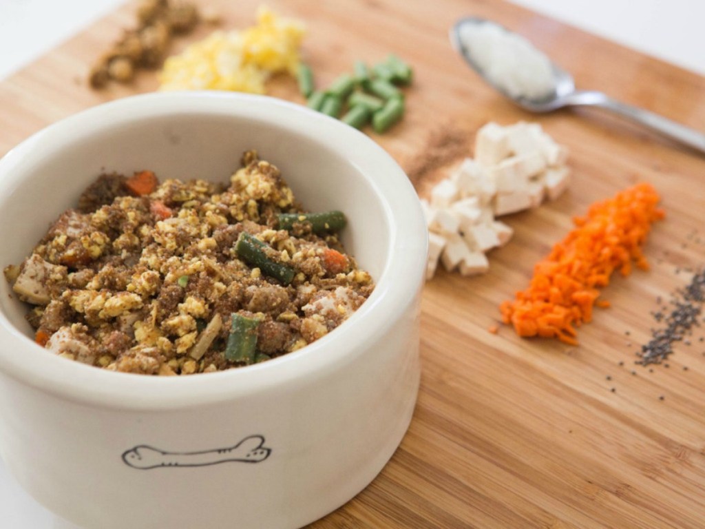 close up of bowl with chopped fresh dog food ingredients in background