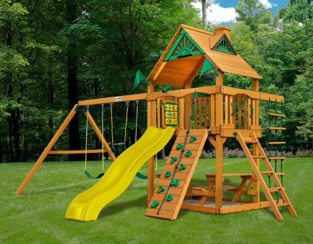 wooden play set with yellow slide and rock wall outside in grass