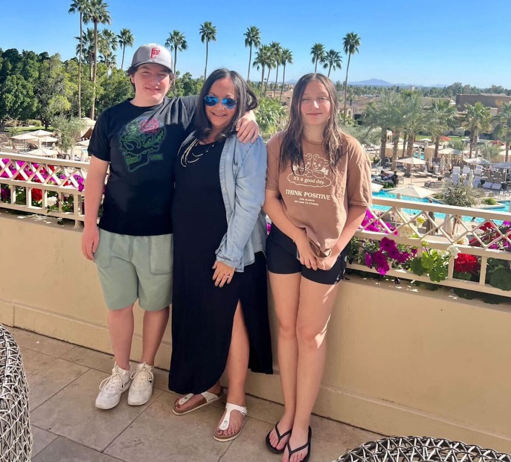 two teens and woman standing on balcony in sun with palm trees