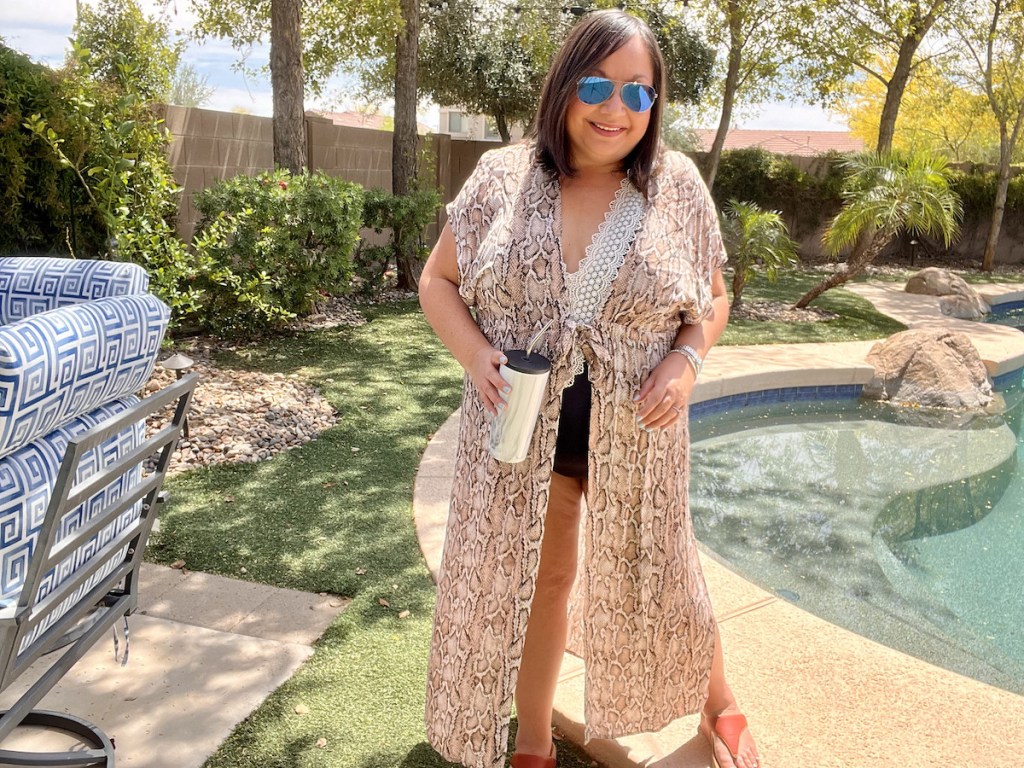 woman standing by pool wearing bathing suit cover up and sunglasses
