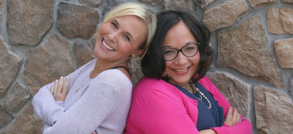 two woman standing back to back in front of stone wall