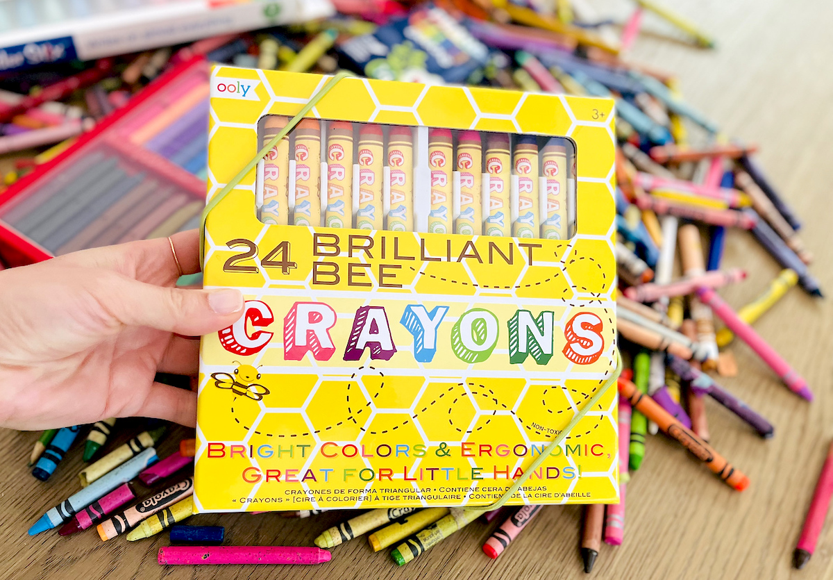 hand holding box of brilliant bee brand crayons on wood table with various crayons spread out