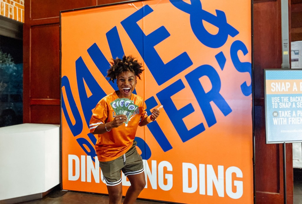 woman standing in front of dave and busters sports bar sign holding paper dollars