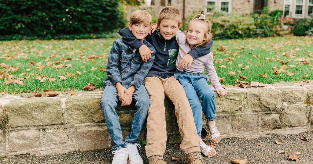 three kids sitting on stone wall outside of house