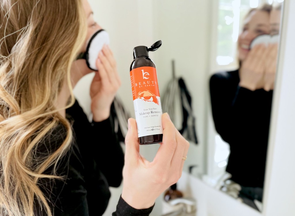 woman looking into mirror washing face while holding bottle of makeup remover