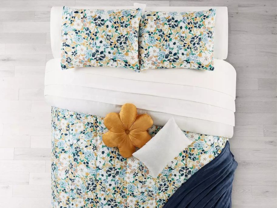 A bedroom with a flowery comforter, flower pillow, and blanket on the bed