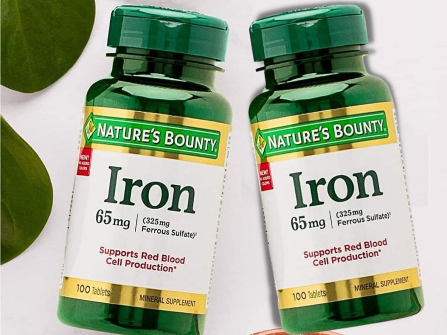 two Natures Bounty Iron Supplements 65 mg 2 Bottles next to leaves on a light grey background