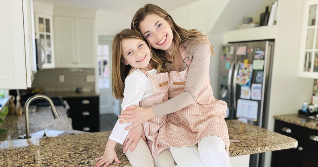 girl and mommy sitting on kitchen countertop wearing matching mommy and me aprons